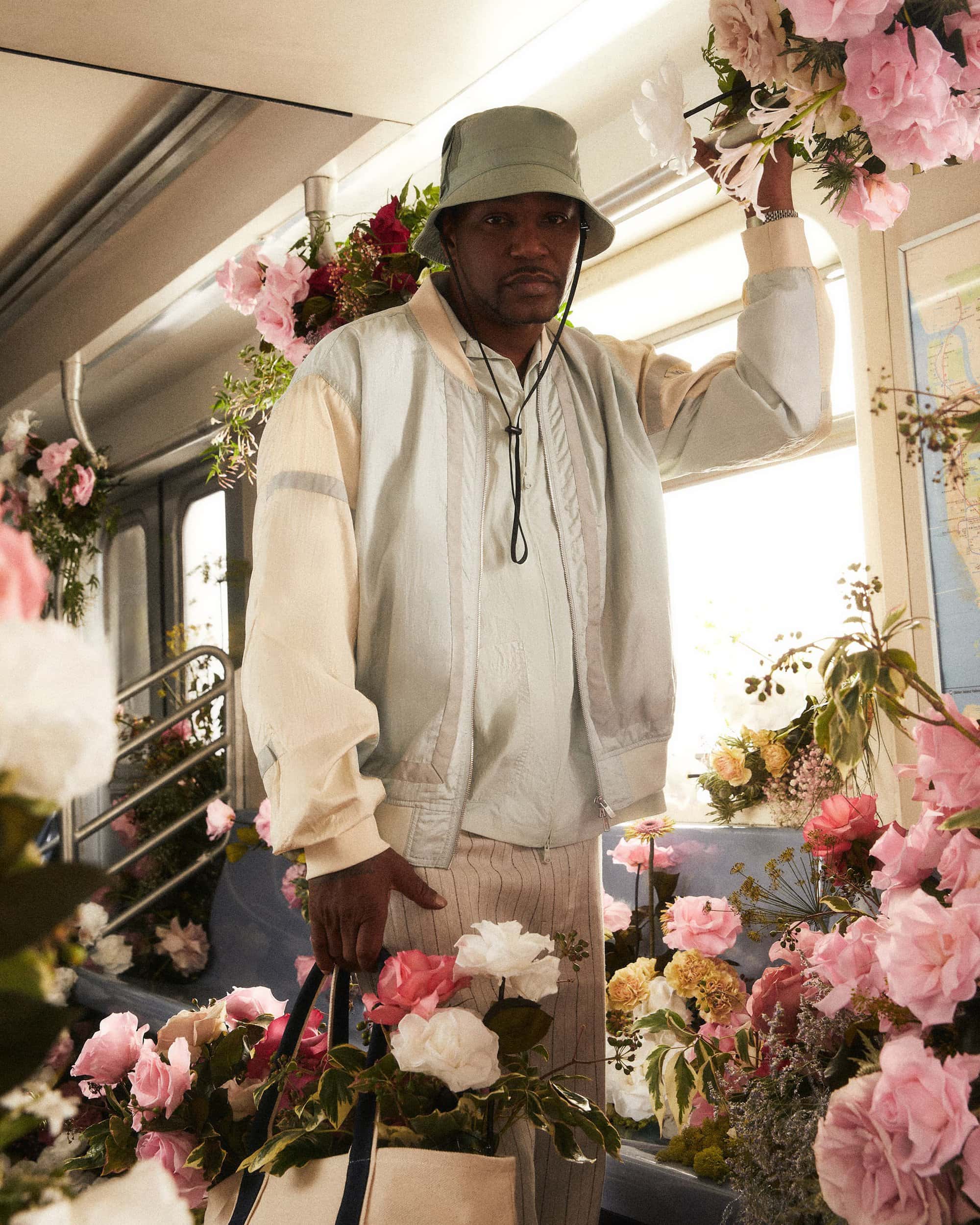 Man in bucket hat on a subway car filled with pink flowers, holding a tote bag of flowers, wearing a pale-blue jacket and striped pants.