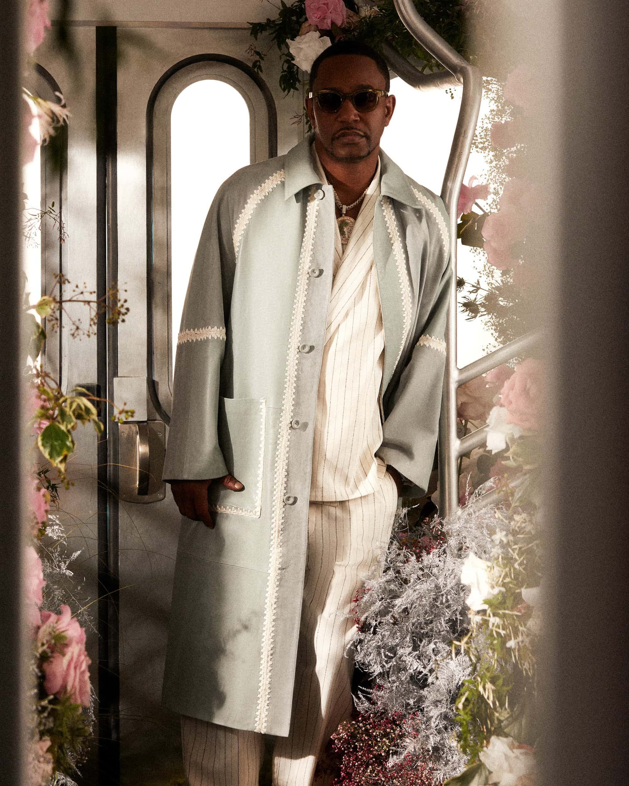 Black man wearing a light green coat with white trim and pinstripe shirt, standing between floral arrangements in a doorway.
