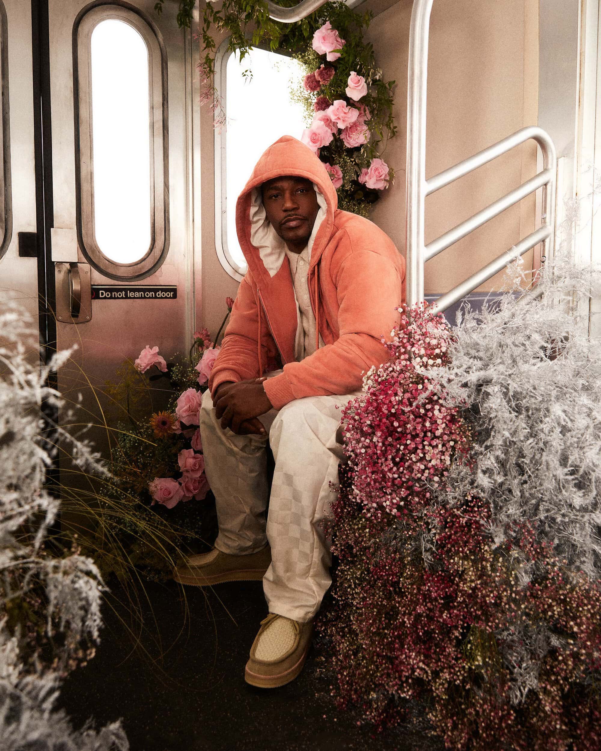 Man in an orange hooded jacket poses sitting in what appears to be a subway car adorned with pink and white flowers and greenery.