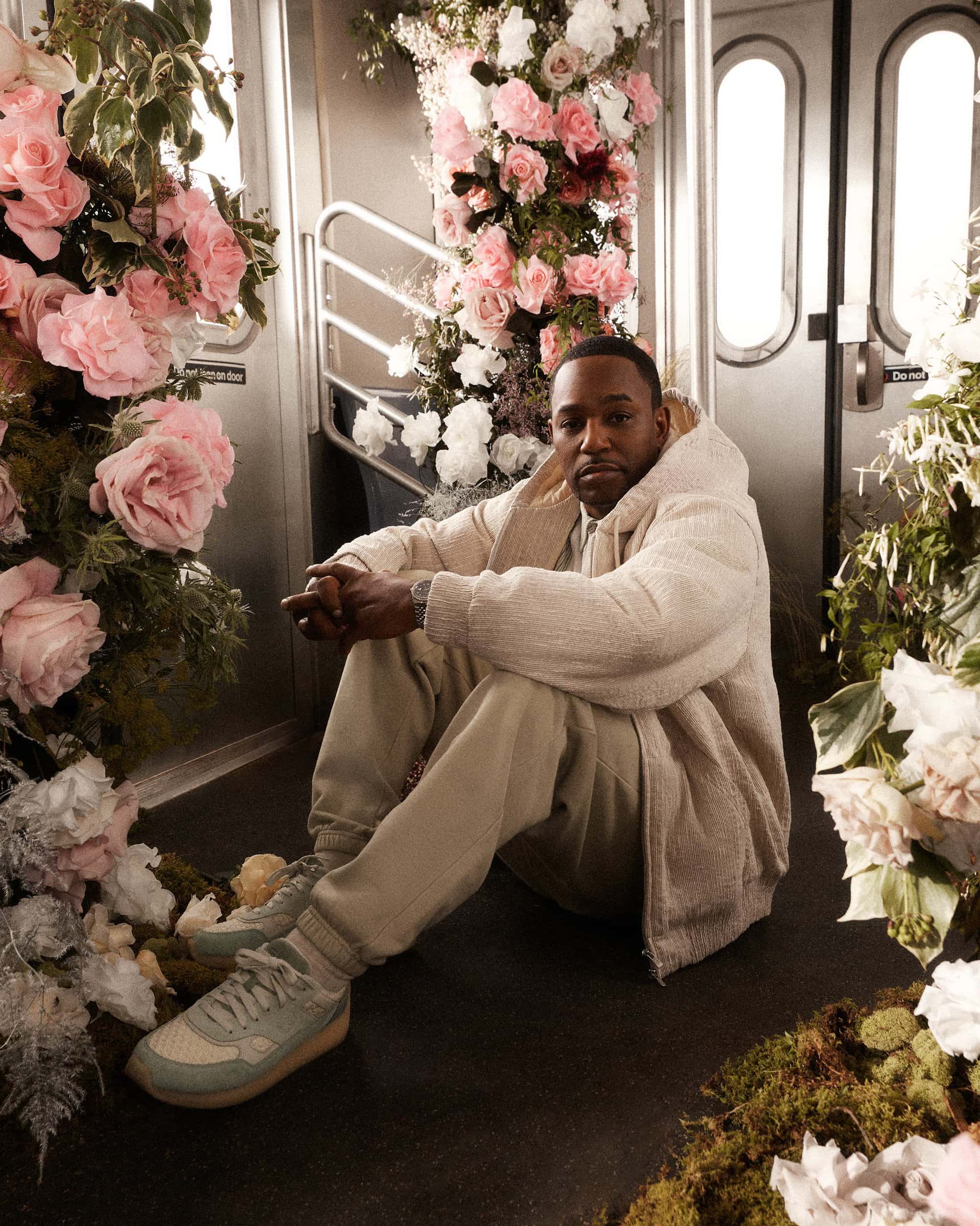 Adult male in a hoodie and sweatpants sits in a subway car decorated with pink and white flowers and green foliage.