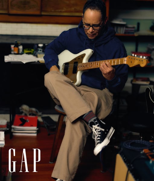 Adult male playing a white electric guitar indoors while sitting on a stool, wearing a blue hooded sweatshirt and khaki pants with the GAP logo.