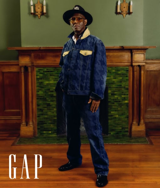 Full-length portrait of a mature Black man in denim jacket and hat, standing in front of a fireplace, promoting the GAP brand.