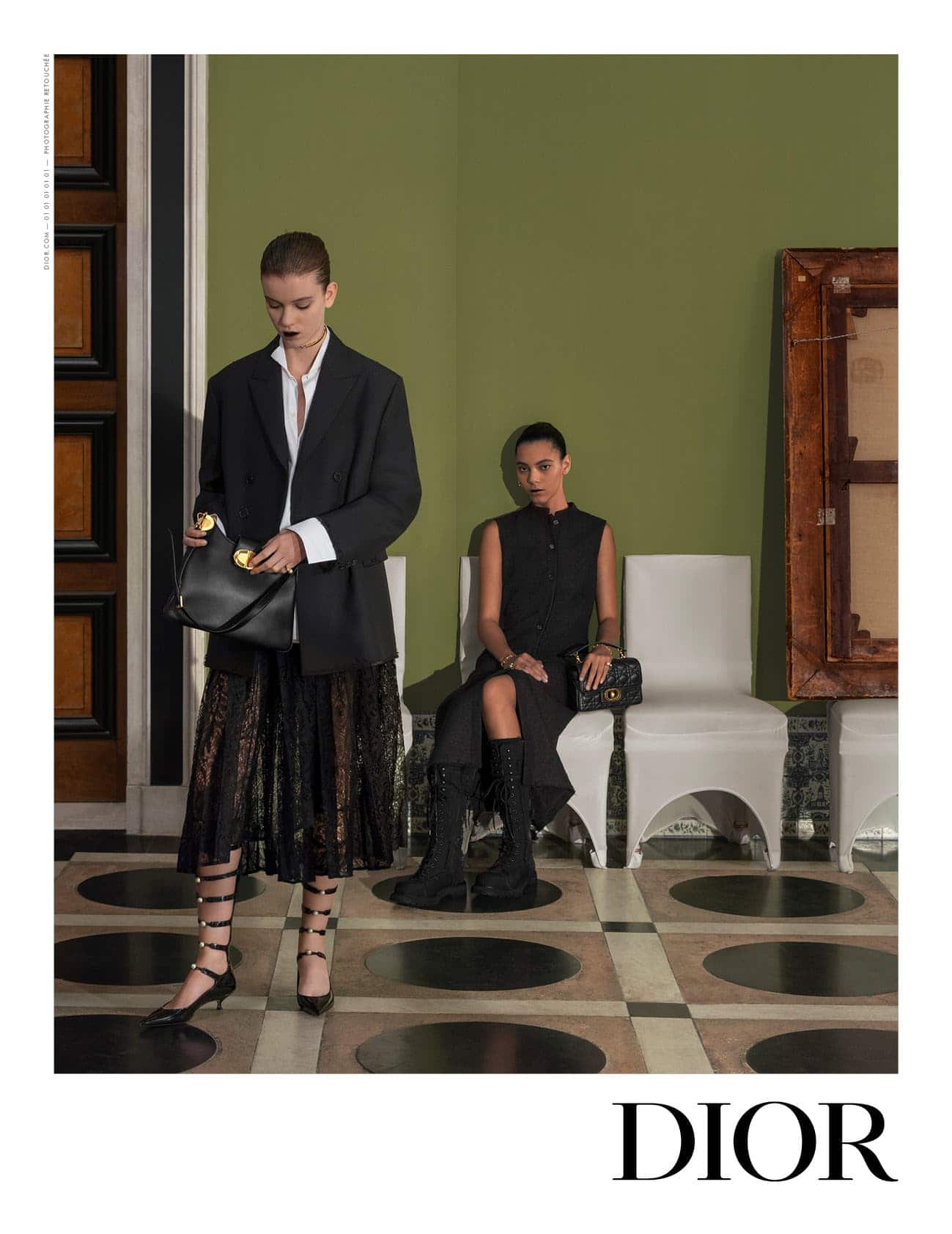 Two fashion models in a photo studio. One woman stands holding a black purse, while the other sits on a white chair.