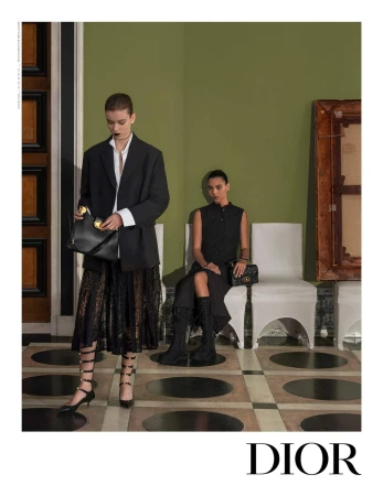 Two fashion models in a photo studio. One woman stands holding a black purse, while the other sits on a white chair.