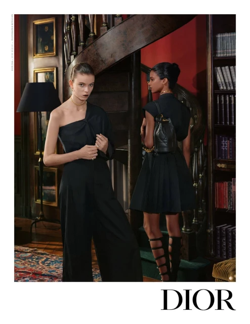 Two fashion models wearing elegant black dresses pose in an interior with a staircase and bookshelves, showcasing a luxury fashion brand.