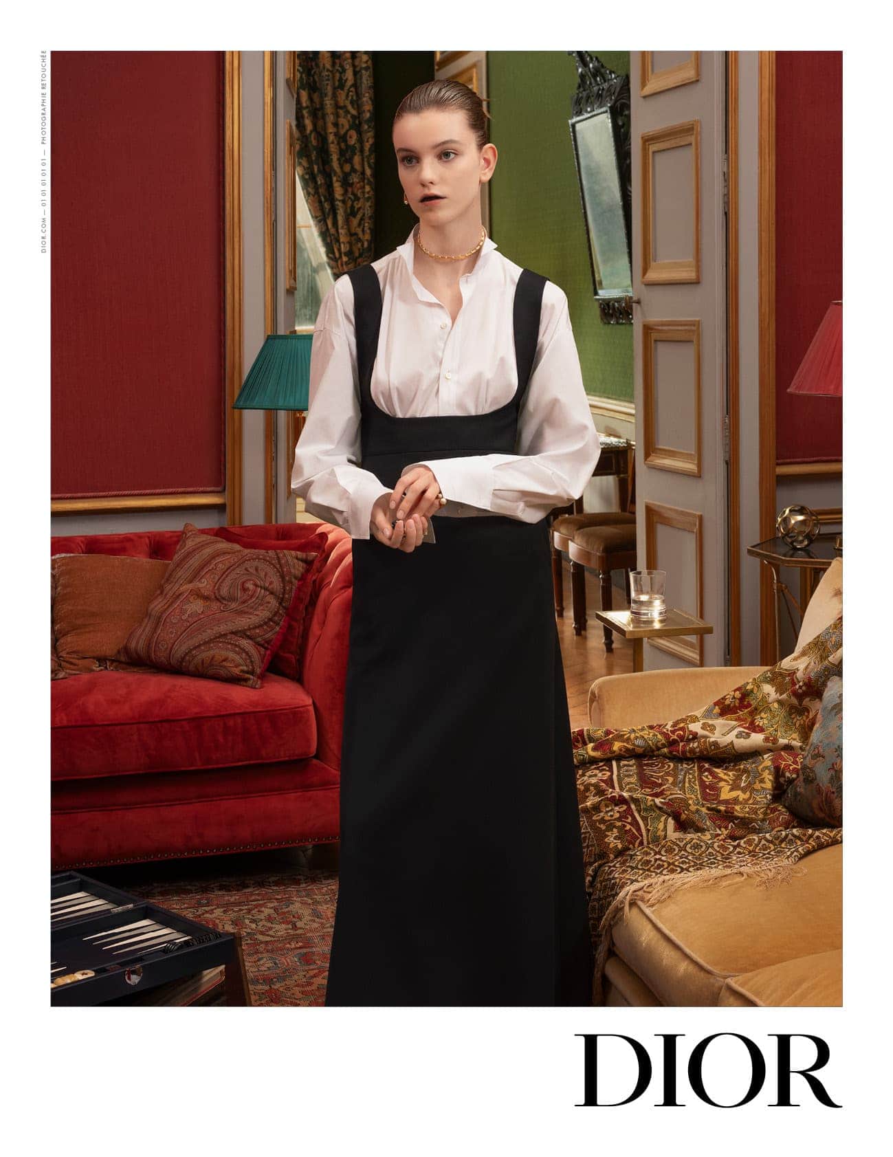 Fashion shoot of a young adult female wearing a black dress over a white blouse in an ornate interior setting with classic furnishings.