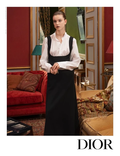 Fashion shoot of a young adult female wearing a black dress over a white blouse in an ornate interior setting with classic furnishings.