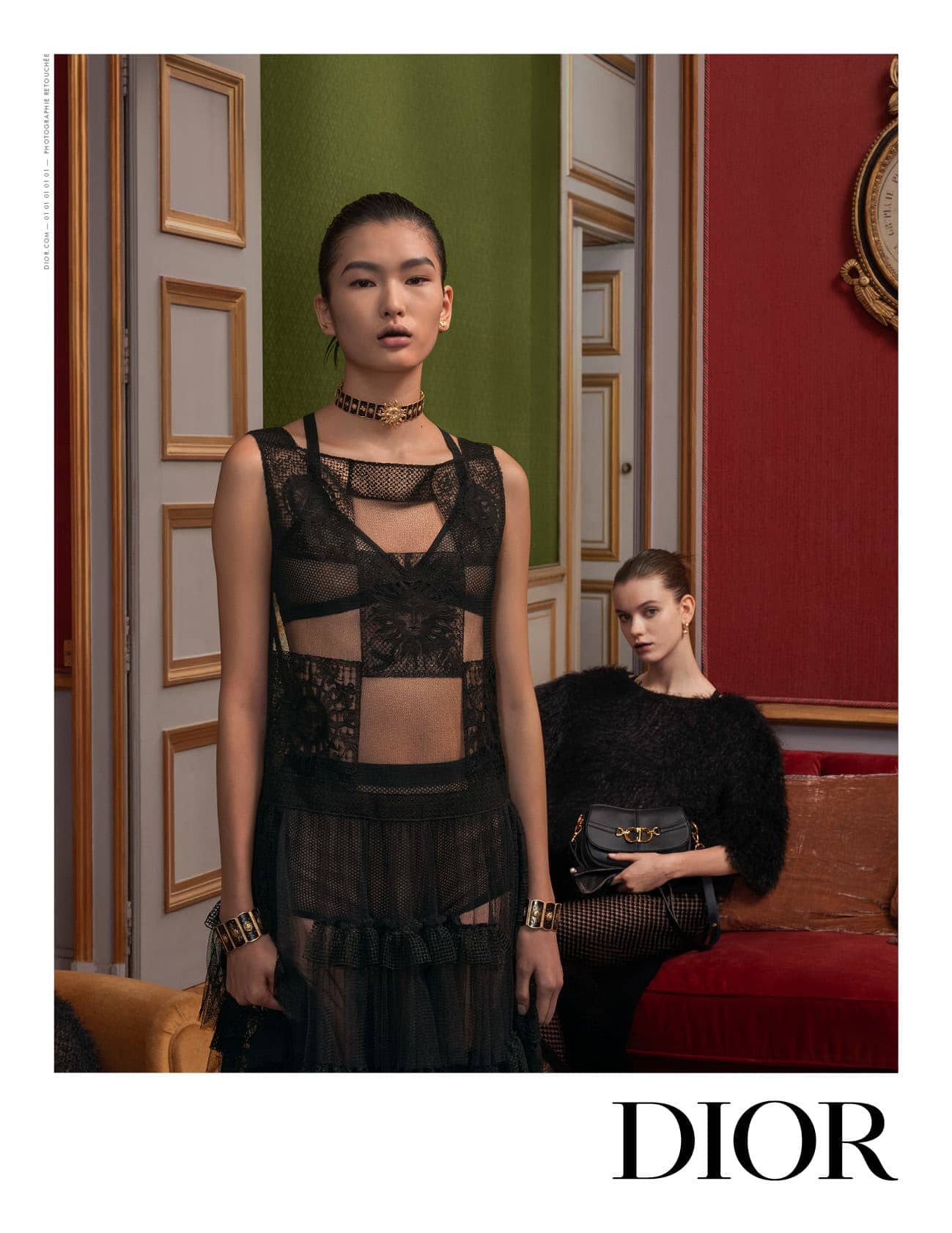 Two fashion models pose indoors in black lace dresses, one standing and one seated, showcasing a black purse against a red wall.