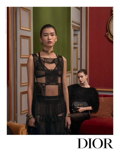 Two fashion models pose indoors in black lace dresses, one standing and one seated, showcasing a black purse against a red wall.
