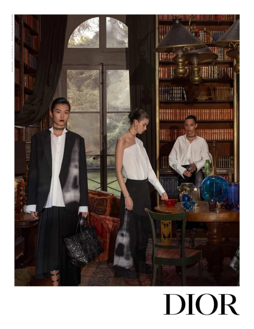 Three fashion models pose in an elegant room with bookshelves, a large window, and decorative objects, showcasing elegant modern designs.