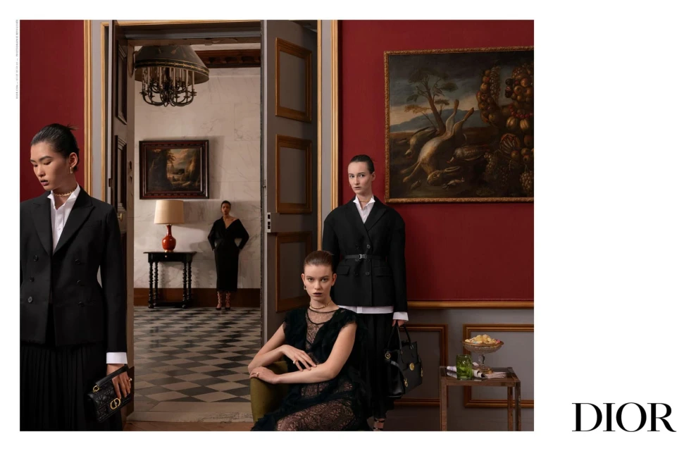 Three women in formal black outfits pose in an ornate room with paintings, an antique lamp, and decor, blending fashion with classic art.
