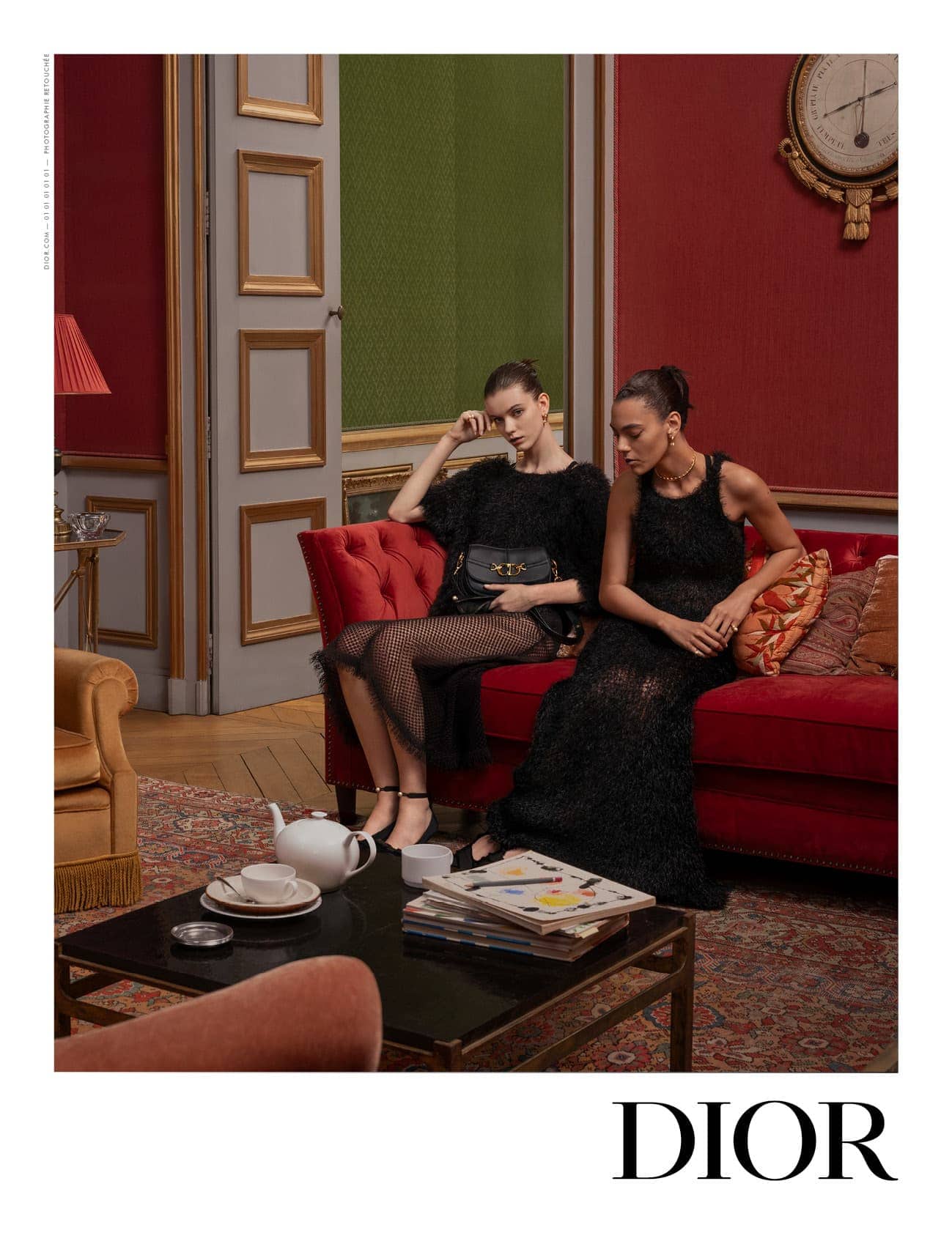 Two fashion models in black dresses pose indoors on a red couch in an editorial shot for Dior, with a tea set on the coffee table.