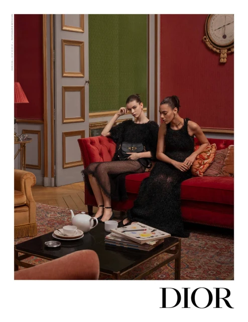 Two fashion models in black dresses pose indoors on a red couch in an editorial shot for Dior, with a tea set on the coffee table.