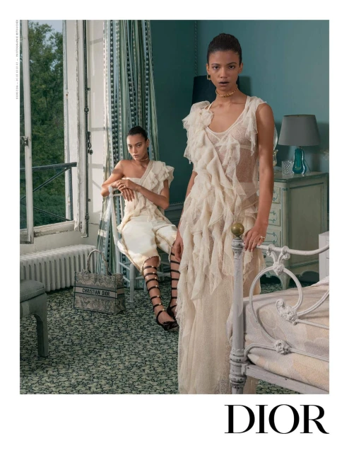 Two female fashion models pose in ruffled off-white dresses near a window. A Dior tote bag sits on the floor. Soft, diffused lighting.