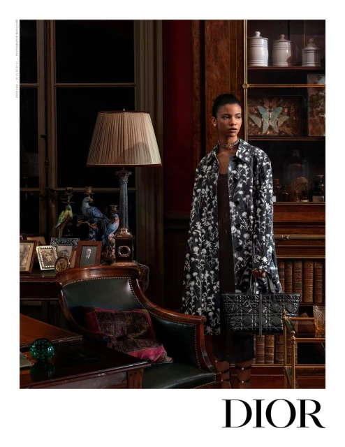 Editorial fashion shot of a Black woman in a patterned coat and black dress, holding a purse, in a vintage-styled room with wood furniture.