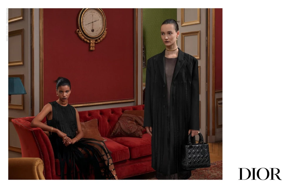 Two women wearing elegant black outfits are staged inside an ornate room with a red couch, antique clock, and luxury handbag by Dior.