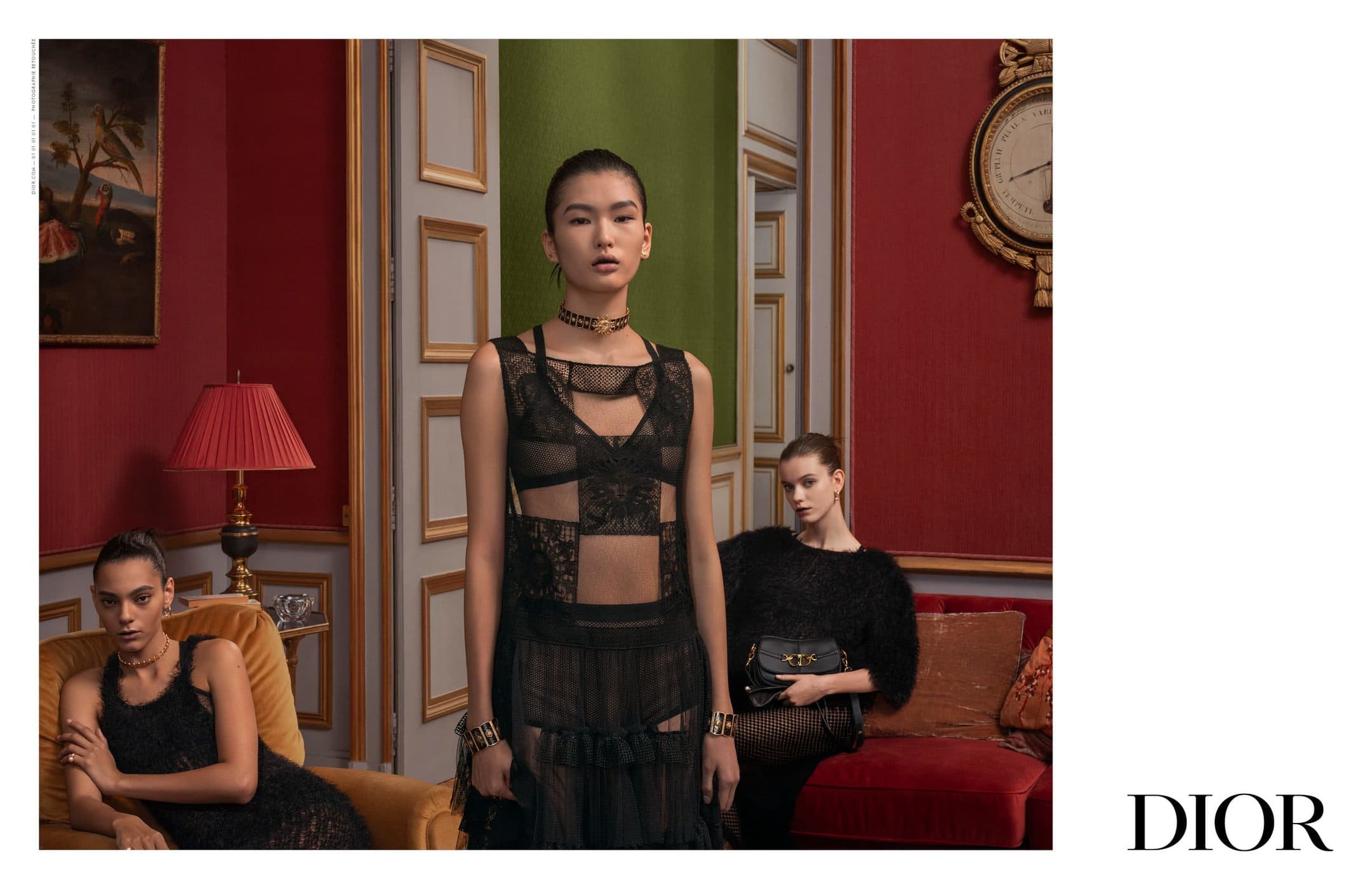 Three fashion models pose indoors against red and green walls with gold trim. The models wear black dresses and hold handbags.