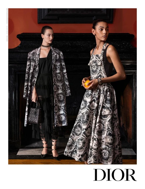 Two women showcasing black and white print dresses in an indoor setting with a fireplace and orange, posing for a fashion advertisement.