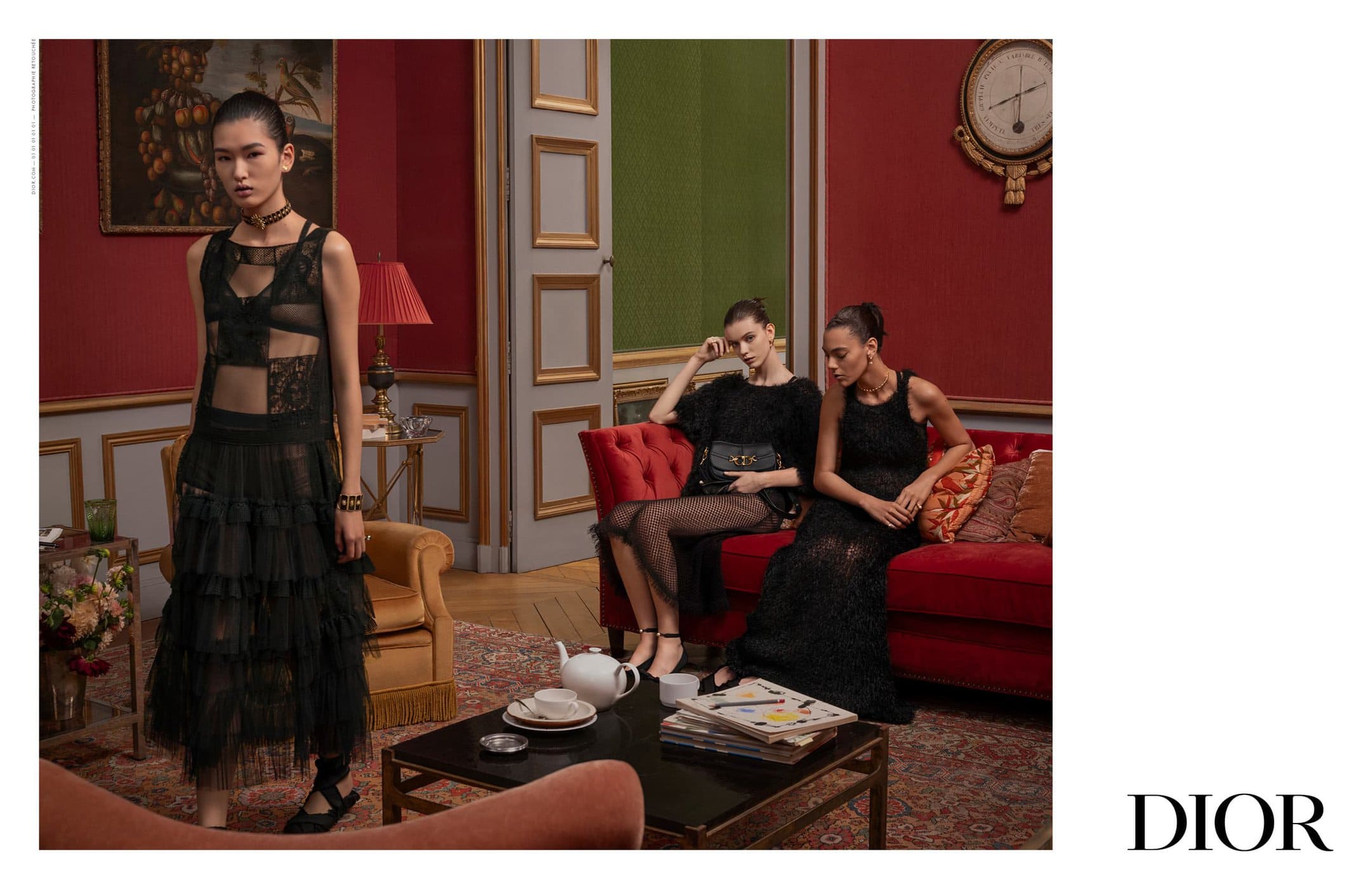 Three women pose in black lace dresses in a vintage-style room with red walls and a red sofa, showcasing high fashion and elegant design.