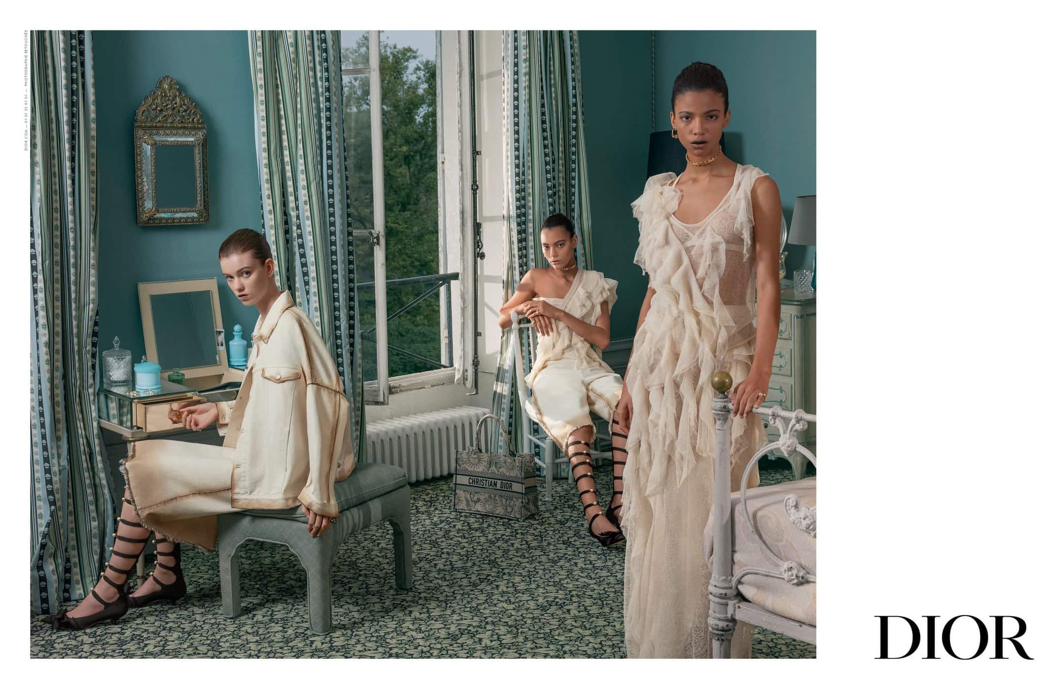 Three fashion models pose in a naturally lit interior wearing cream-colored, layered dresses. The setting includes a window, dressing table, and soft greens.