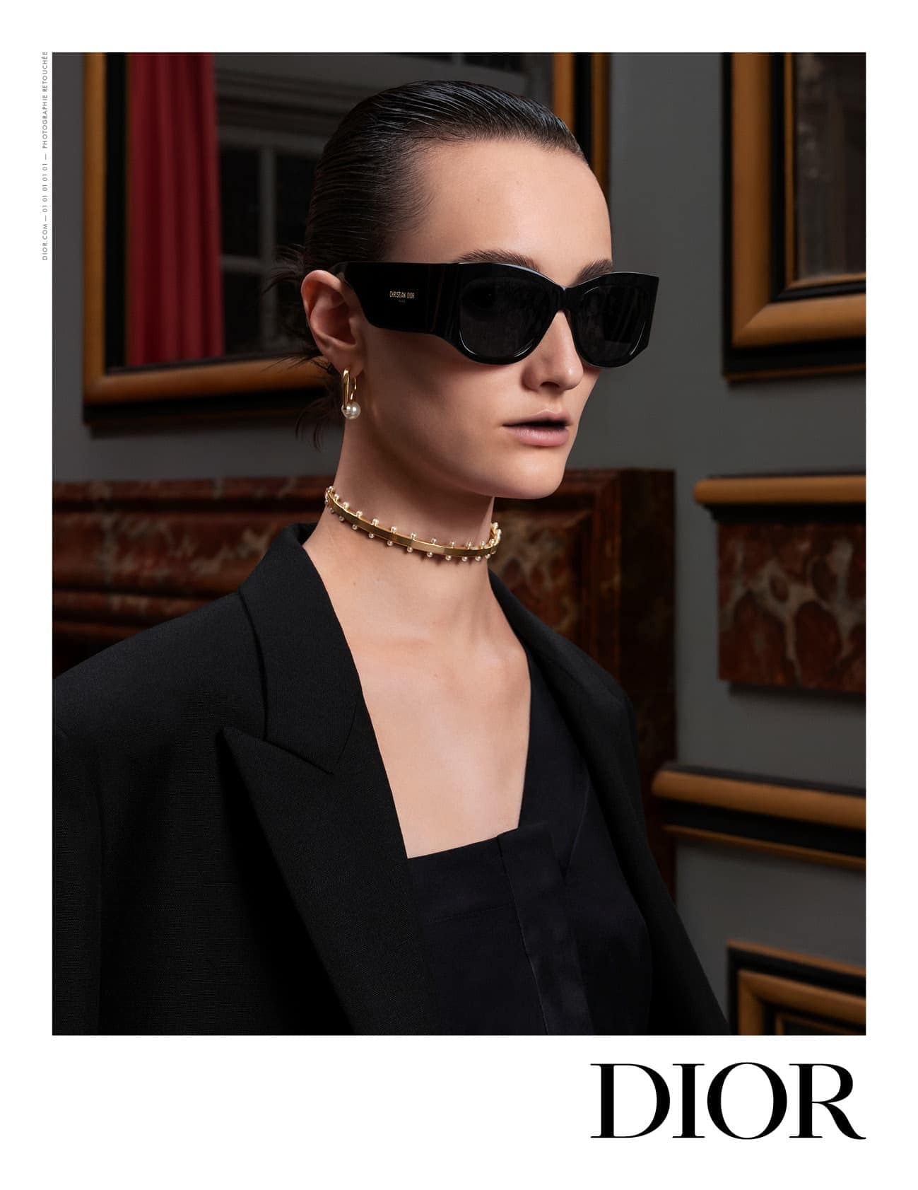 Elegant woman poses indoors wearing a black blazer, black sunglasses, and a beaded choker necklace, in a fashion editorial shot.