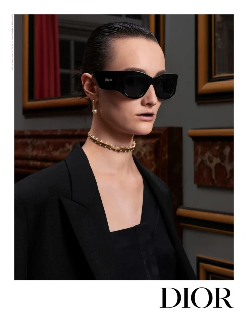 Elegant woman poses indoors wearing a black blazer, black sunglasses, and a beaded choker necklace, in a fashion editorial shot.