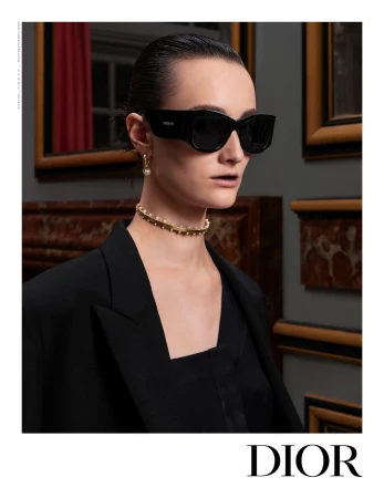 Elegant woman poses indoors wearing a black blazer, black sunglasses, and a beaded choker necklace, in a fashion editorial shot.