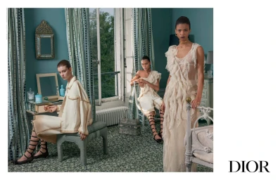Three female models in cream designer clothing inside a teal bedroom with patterned curtains and a window overlooking trees.