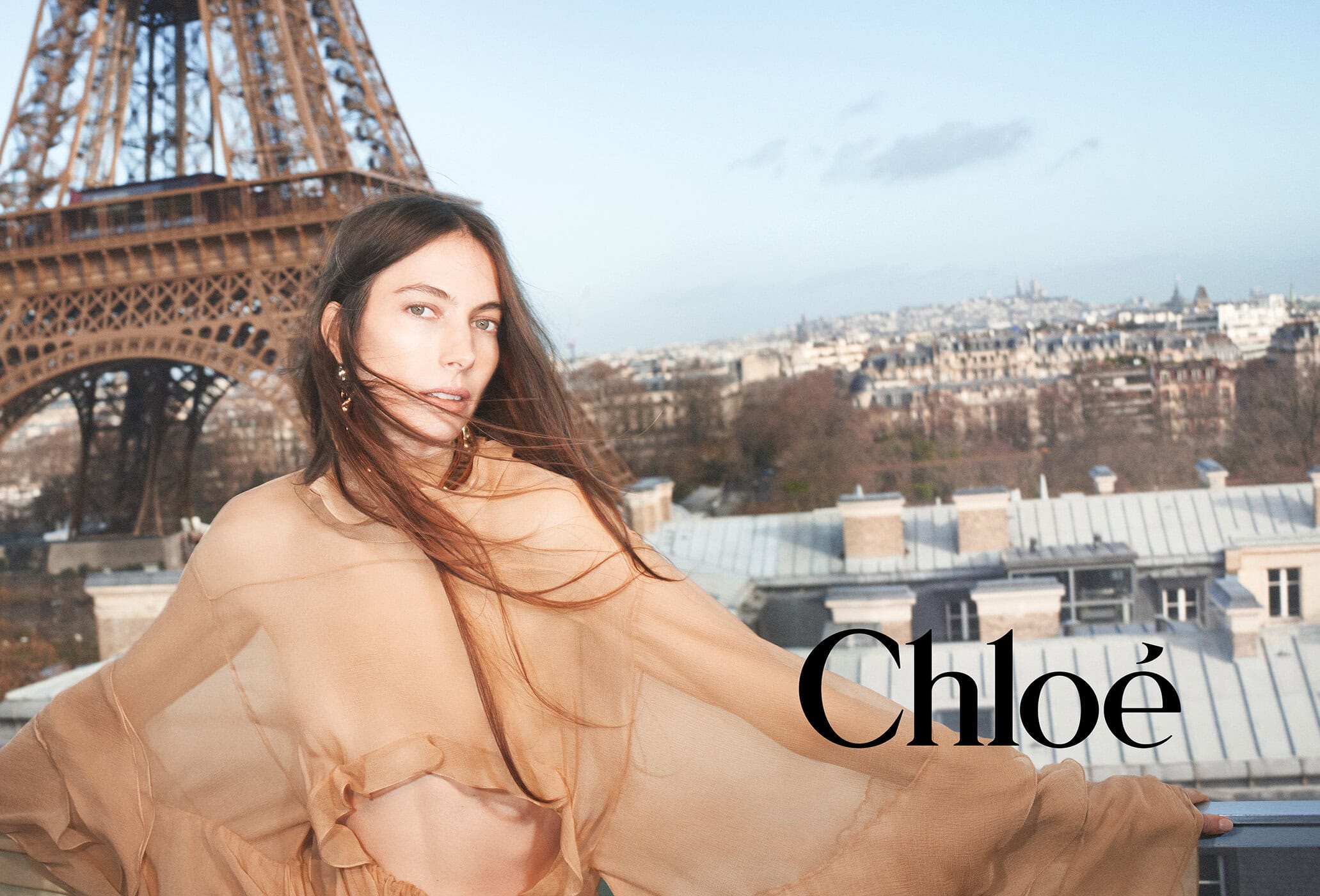 Fashion portrait of a woman in a tan sheer top, posing on a rooftop with the Eiffel Tower and a Parisian cityscape in the background.