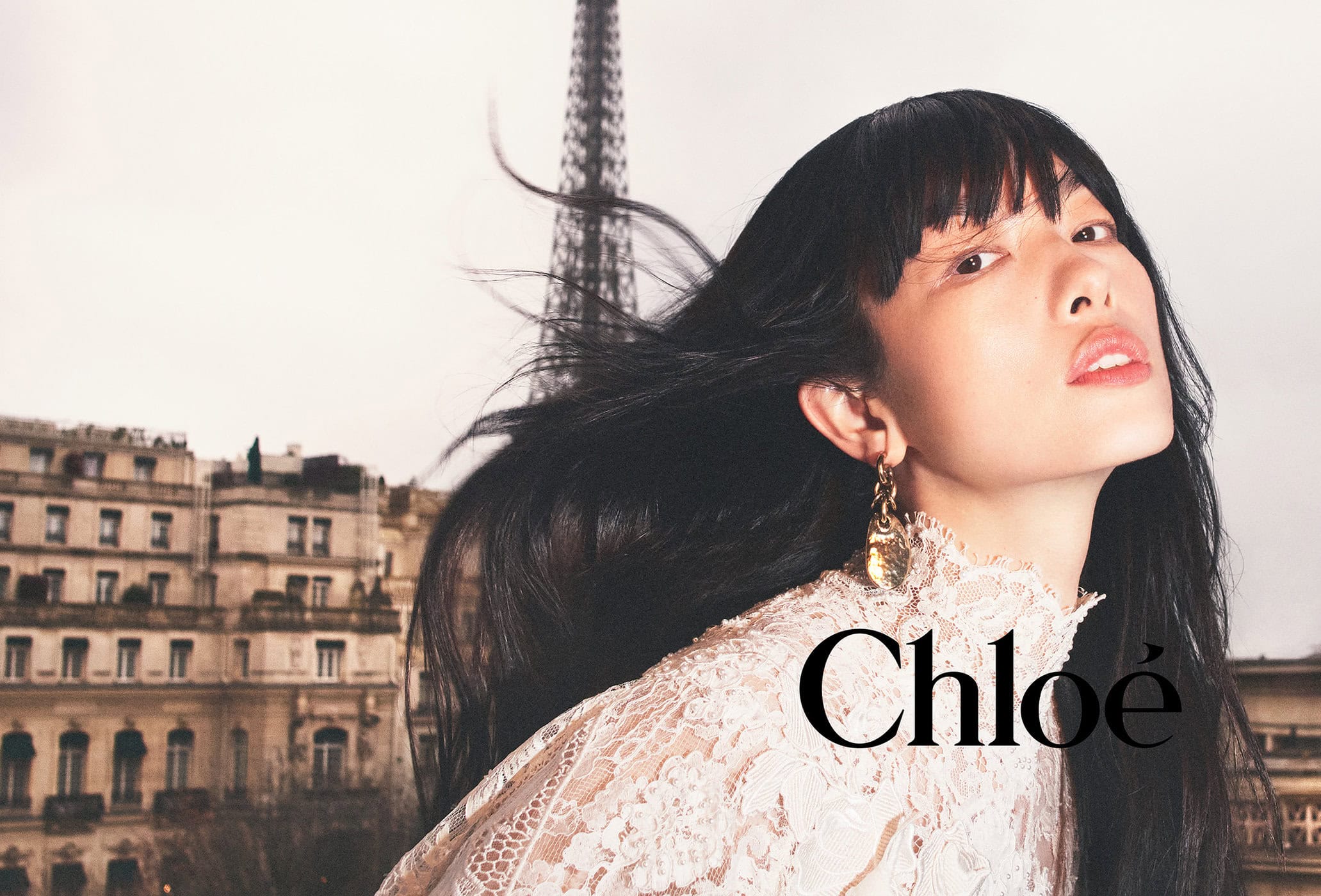 Fashion shot of a woman with black hair wearing a lace top, posed against a Paris cityscape, with the Eiffel Tower in the distance.