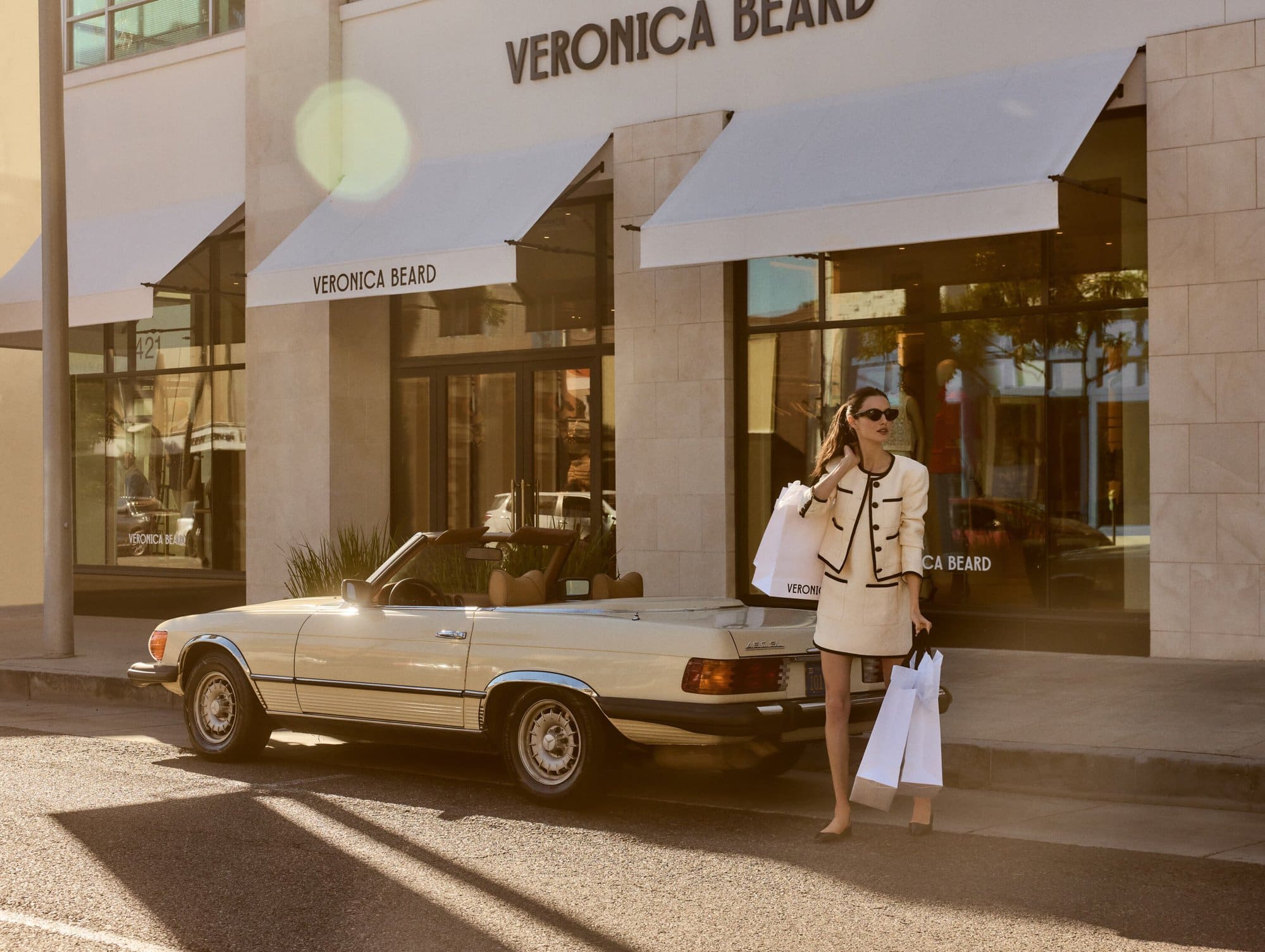 A woman in a cream skirt suit stands outside a Veronica Beard store with a cream convertible car and shopping bags in hand.