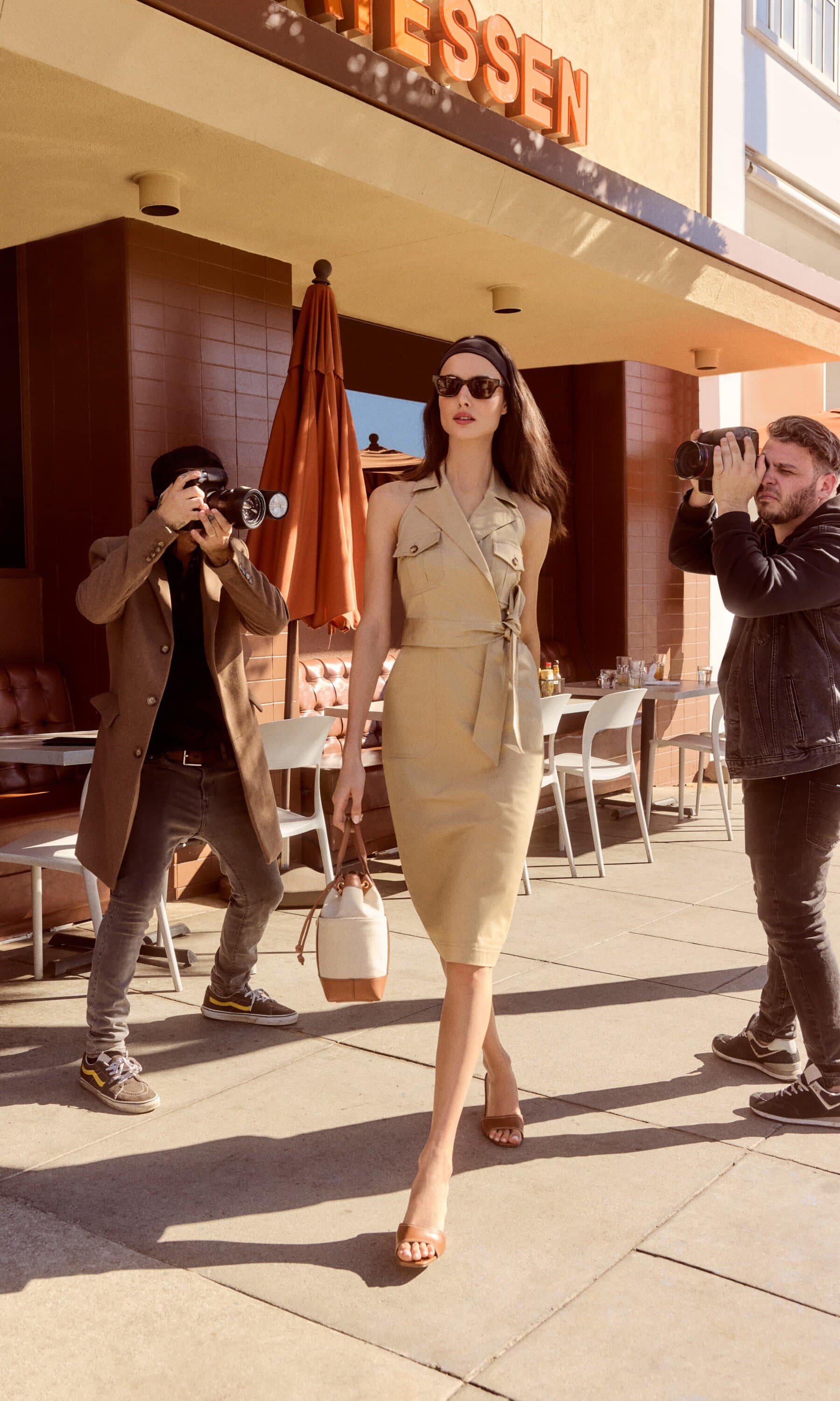 A woman in a beige dress walks between two photographers outside a building with a restaurant during daytime, fashion shoot concept.