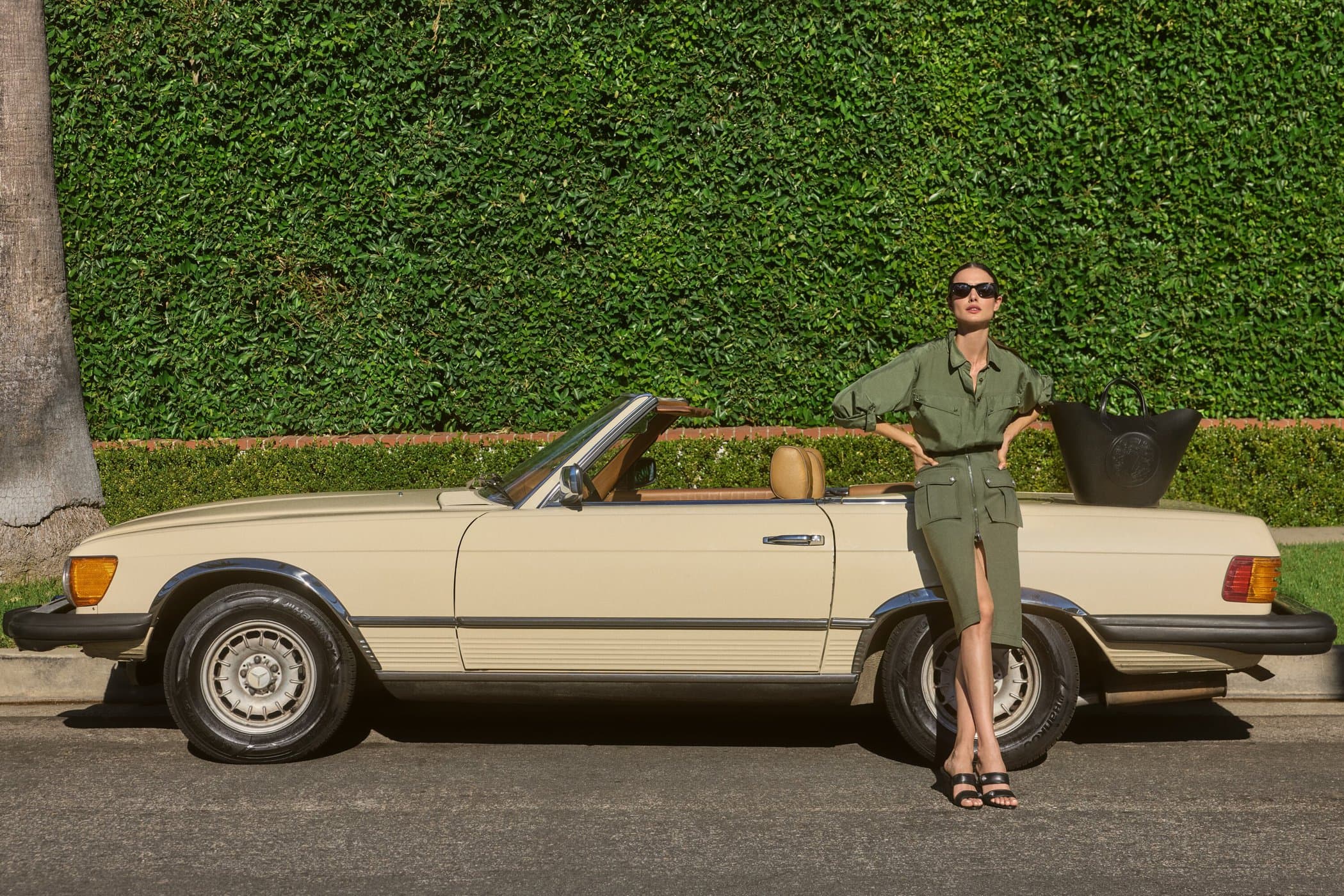 Adult woman posing with a classic convertible car in front of a lush green hedge, wearing sunglasses and a green utility-style outfit.