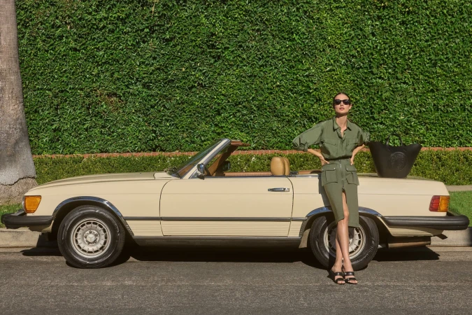 Adult woman posing with a classic convertible car in front of a lush green hedge, wearing sunglasses and a green utility-style outfit.