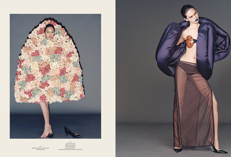 Two women pose in avant-garde fashion against a gray studio backdrop; one wears a floral structure and the other a jacket and skirt.