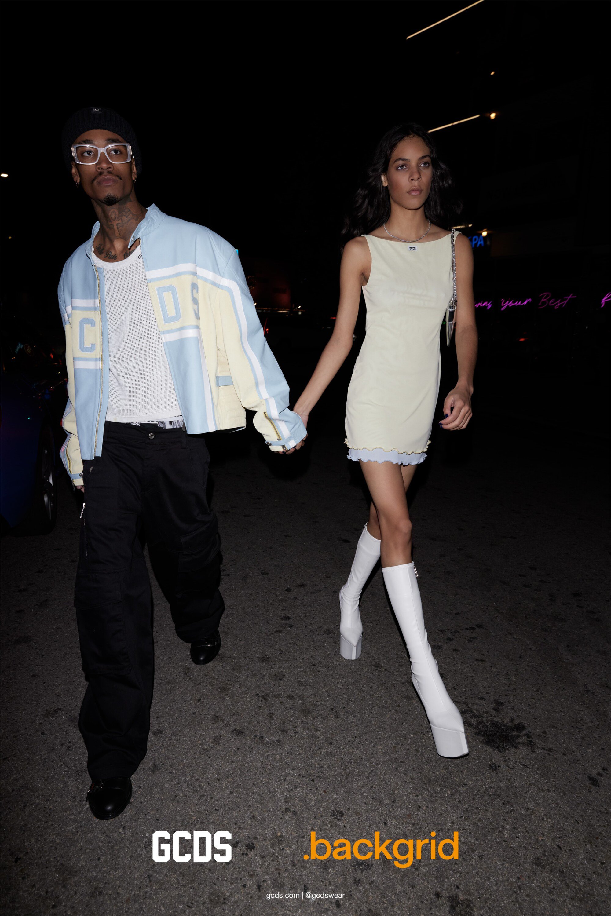 A man and woman hold hands on a city street at night. The woman wears a white dress and boots while the man is in casual attire.