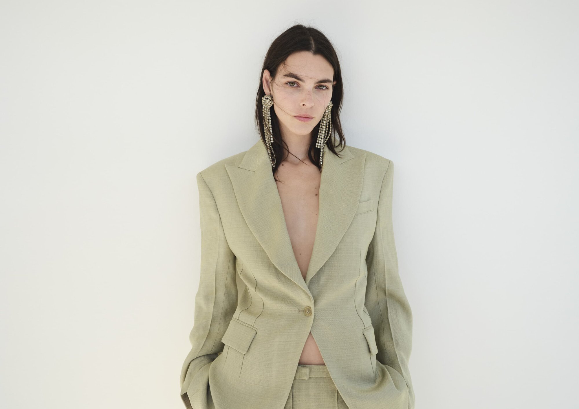 Portrait of a woman wearing a beige blazer and crystal earrings against a white background in a fashion style pose.