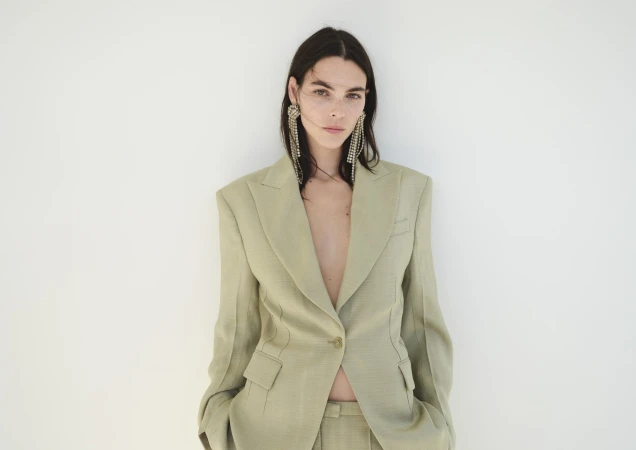 Portrait of a woman wearing a beige blazer and crystal earrings against a white background in a fashion style pose.