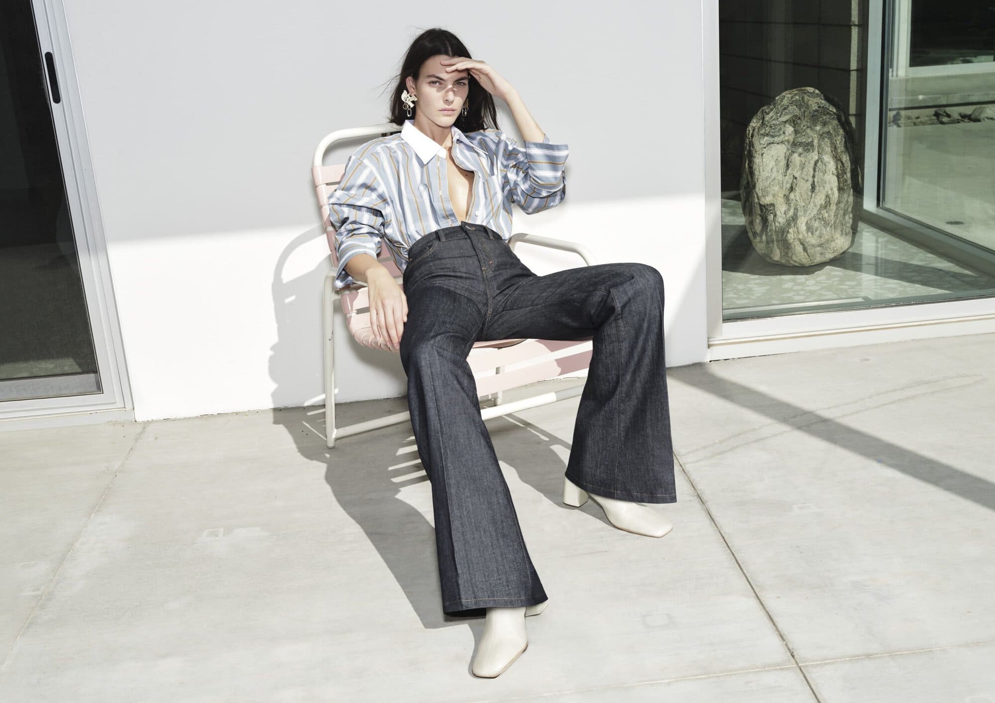 Woman in striped blouse and denim jeans sitting on a chair outdoors in sunlight, with a modern stone sculpture in the background.