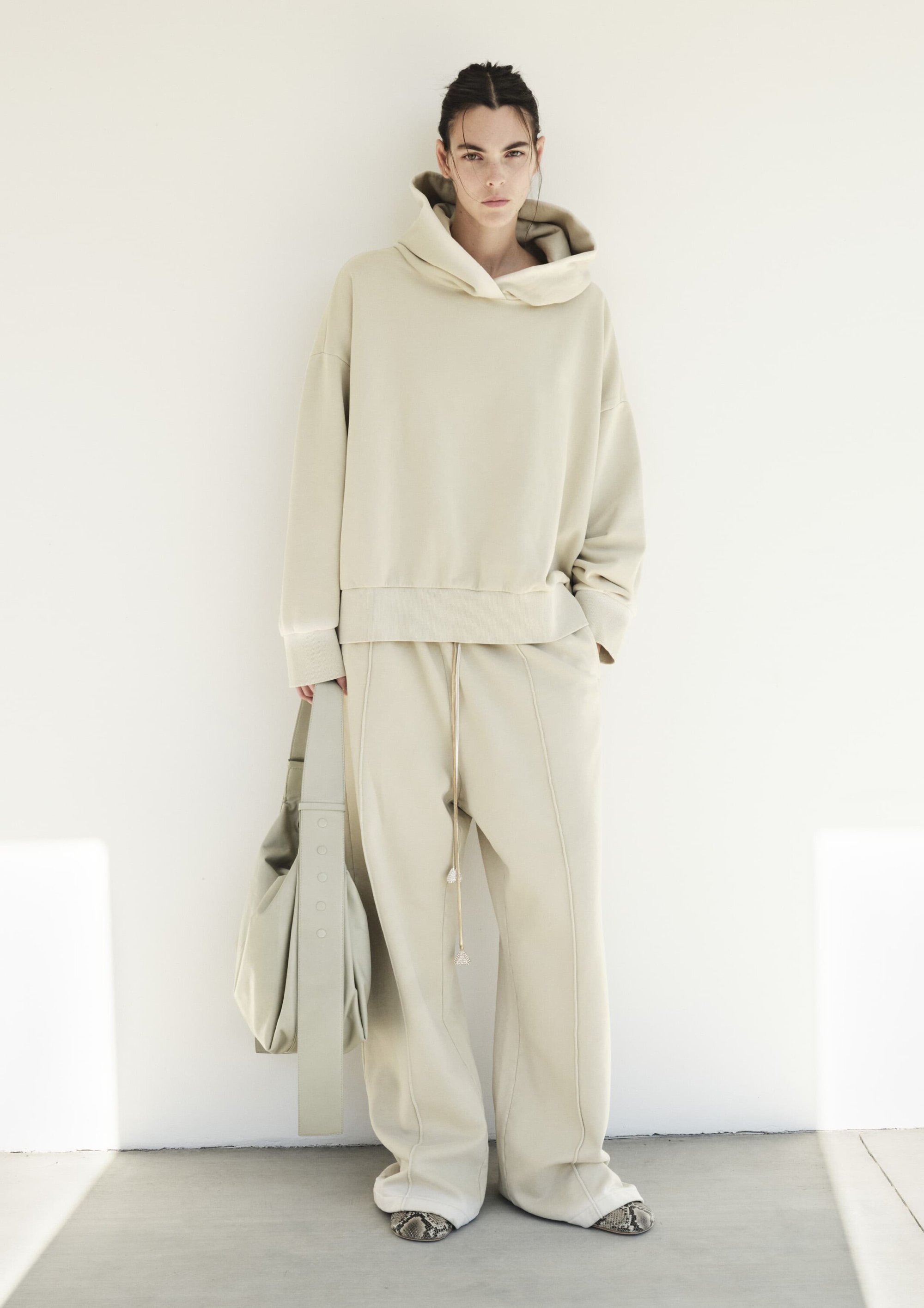 A woman in a cream-colored hoodie and pants poses against a white wall, holding a matching bag, showcasing neutral tones.