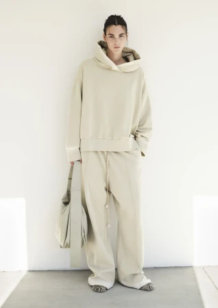 A woman in a cream-colored hoodie and pants poses against a white wall, holding a matching bag, showcasing neutral tones.