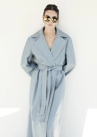 Woman in powder-blue belted trench coat and reflective sunglasses stands indoors against a white wall in a fashion-style studio portrait.