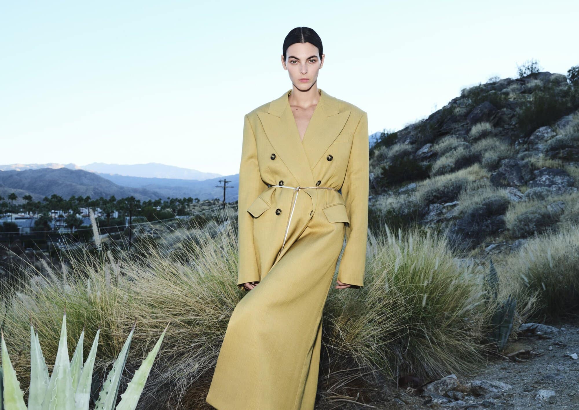 A woman in a long tan coat stands in a dry, grassy landscape with mountains in the background beneath a clear sky, fashion editorial.