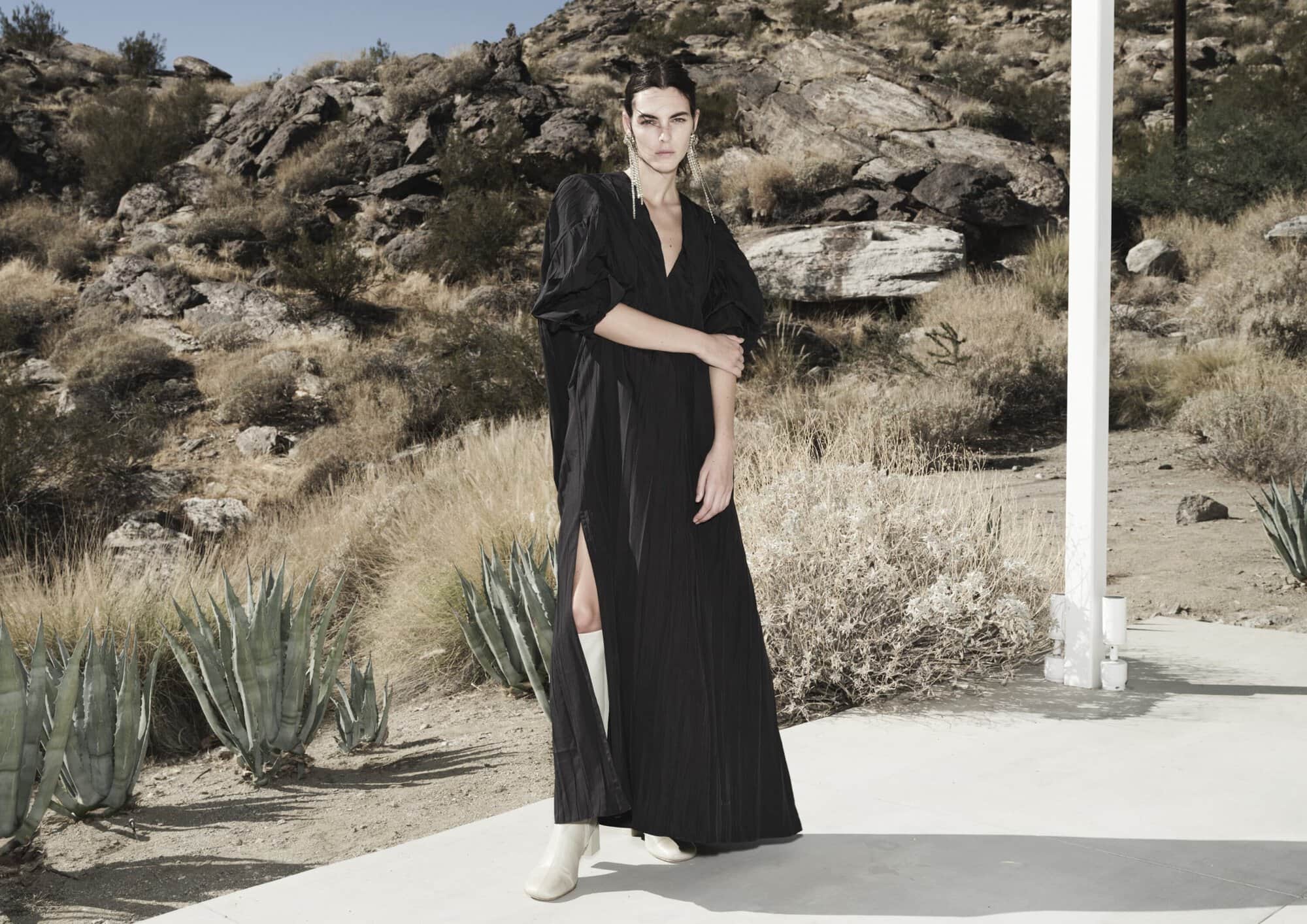 Fashion shot of an adult woman posing in a long black dress with a thigh-high slit, set against a desert landscape with mountains.