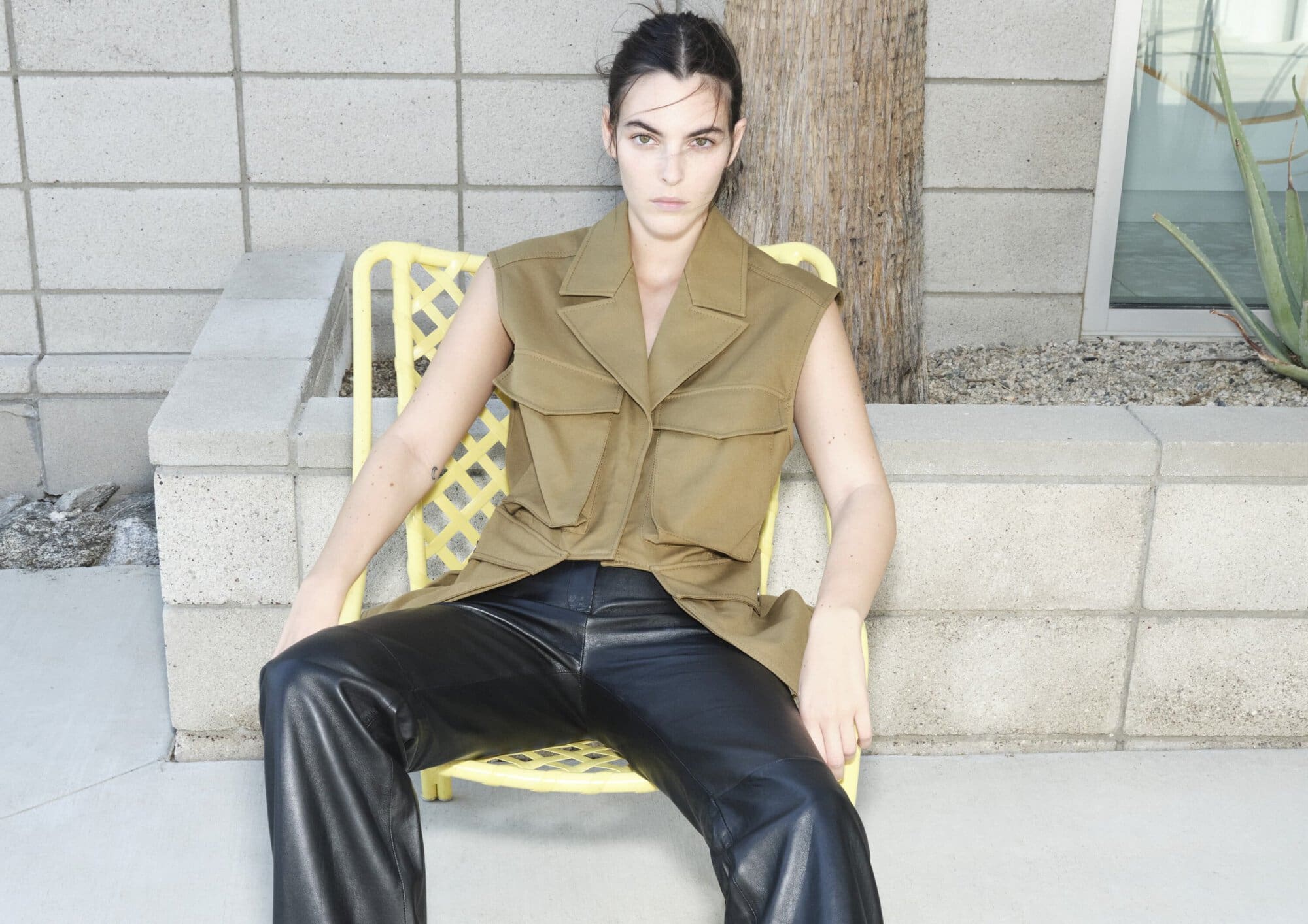 A young adult woman with dark hair poses while seated on a yellow chair wearing a tan vest and black leather pants in an outdoor setting.