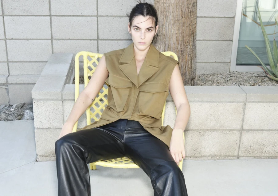 A young adult woman with dark hair poses while seated on a yellow chair wearing a tan vest and black leather pants in an outdoor setting.