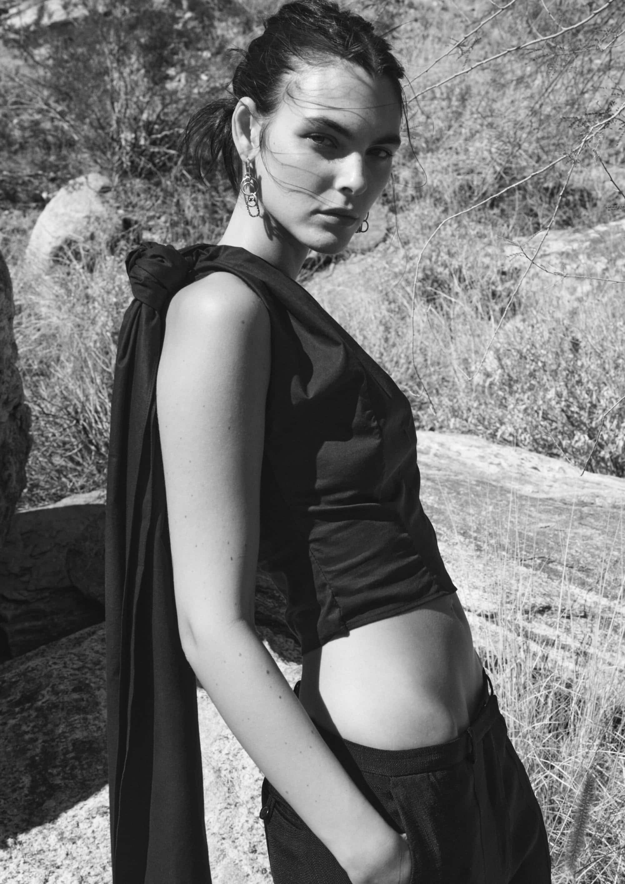Monochrome portrait of a young woman with fair skin in a sleeveless top and pants, with a backdrop of rocks and foliage.