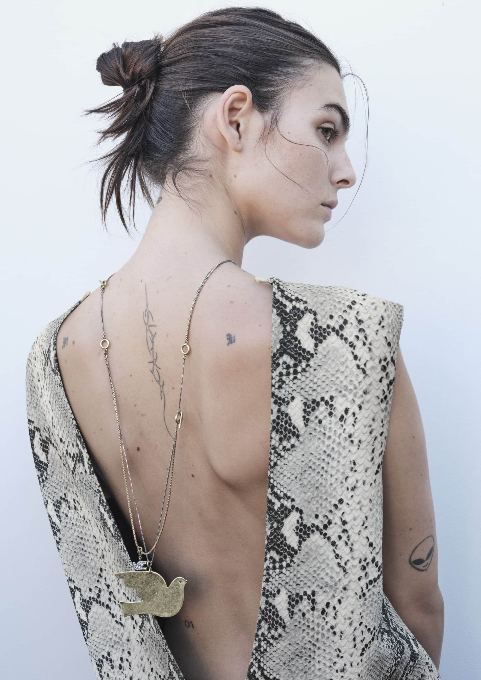Woman with messy bun wears snake-print low-back dress and pendant necklace, showcasing back tattoos in a minimalist studio setting.