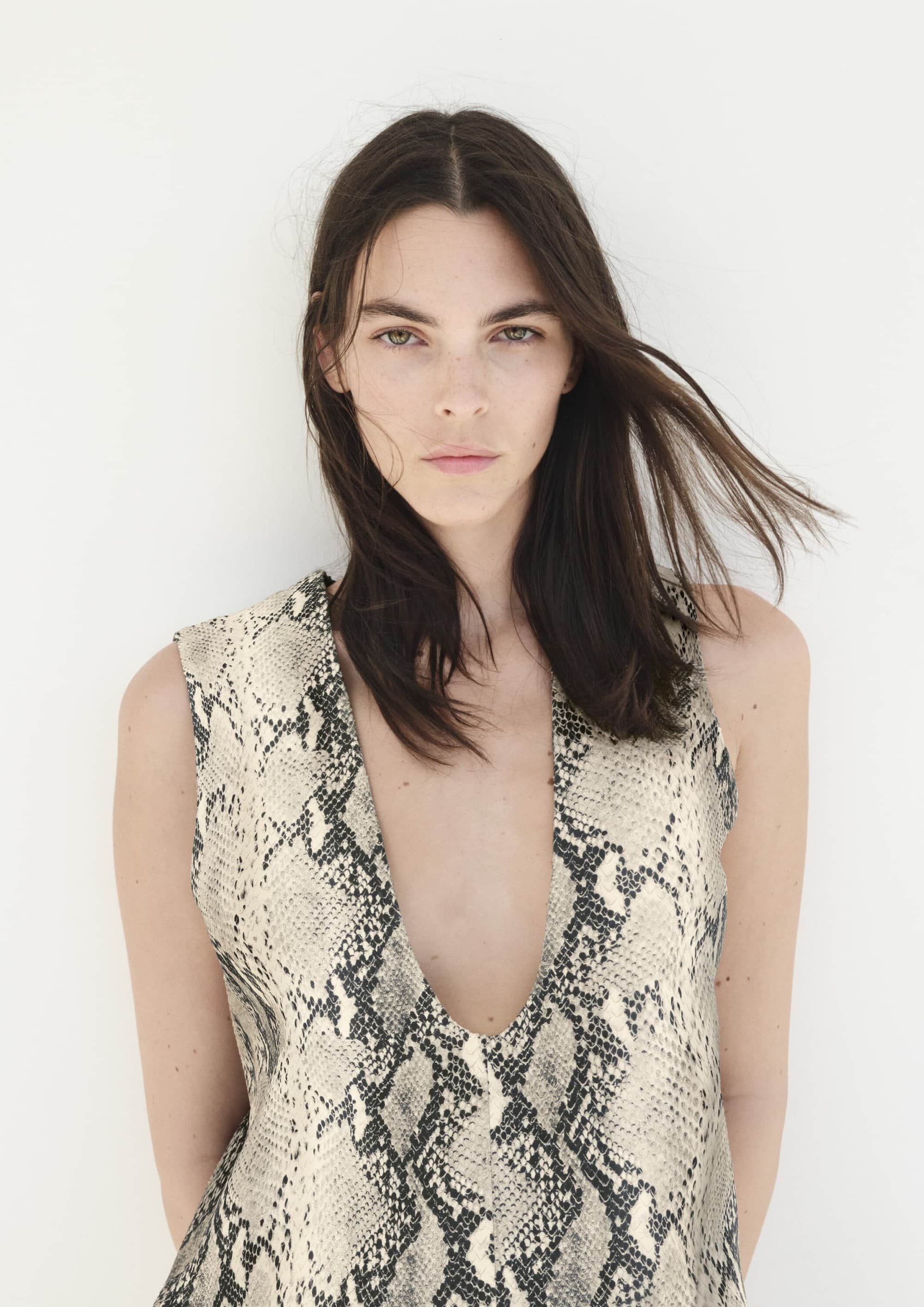 Brunette woman posing in snakeskin-patterned top against a white backdrop, portrait-style fashion shot with natural lighting and neutral tones.