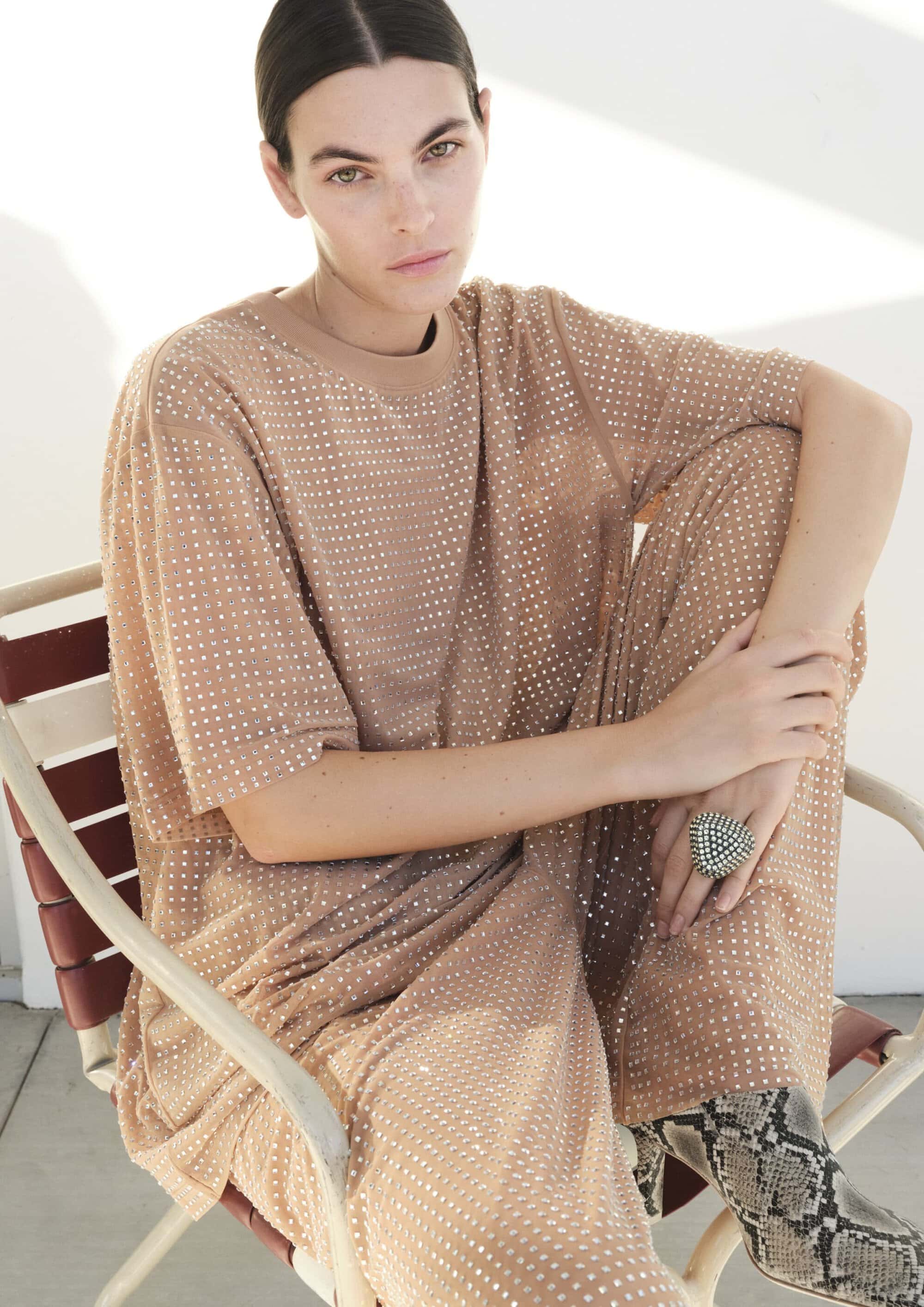 Fashion portrait of a brunette woman in a tan outfit with silver accents, seated on a chair against a neutral background, fashion editorial.